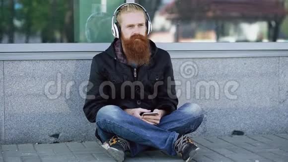 年轻留胡子的嬉皮士带着耳机坐在路上用智能手机听音乐和上网视频的预览图
