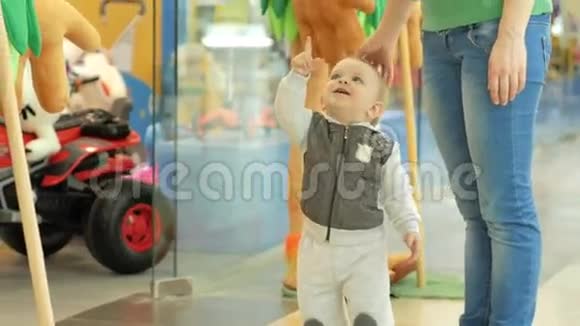 两岁可爱的金发男孩在商店门口的商场里向妈妈展示大软玩具视频的预览图