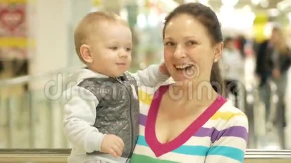 妈妈在商场里抱着一个漂亮的金发儿子在人们去购物的背景下视频的预览图