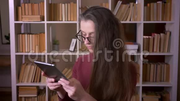 在大学图书馆室内使用平板电脑拍摄戴眼镜的年轻白种人女学生的特写照片视频的预览图