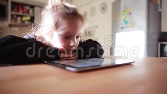 美丽的小女孩坐在桌子旁看着平板电脑上的卡通把头放在手上视频的预览图