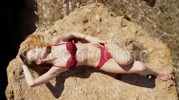 穿红色泳衣的苗条女孩躺在海边的岩石上晒太阳视频的预览图