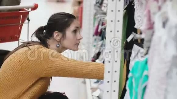 年轻漂亮的女人蹲在超市在橱窗里挑选婴儿服装女的在店里4K视频的预览图