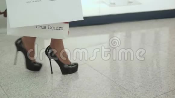 穿着黑色高跟鞋在购物中心散步时一条带着五颜六色购物袋的年轻女子腿特写镜头视频的预览图
