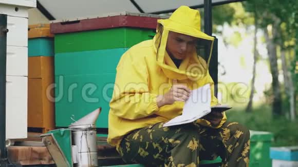 年轻的养蜂人坐在蜂房附近写着记事本检查收获视频的预览图