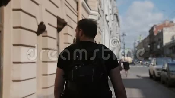 英俊的男人留着胡子探索一座城市穿着黑色背包环顾四周阳光普照慢影稳拍视频的预览图