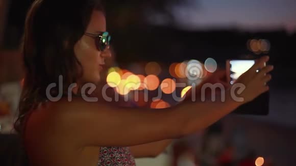 年轻漂亮的黑发女人戴太阳镜摆姿势用智能手机拍自拍照片视频的预览图