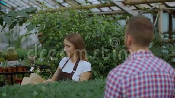 年轻微笑的卖花者在花园中心工作女人给顾客一个购物袋用信用卡付款视频的预览图
