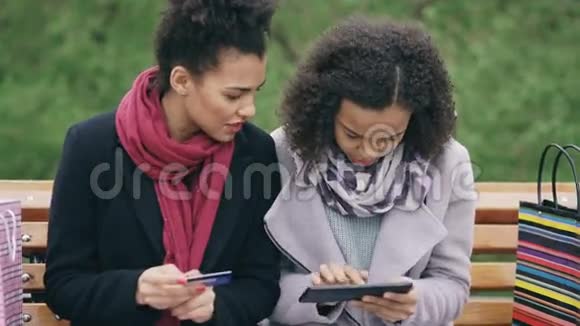 两位漂亮的混血女人她们拿着纸袋坐在长凳上用平板电脑和信用卡上网视频的预览图