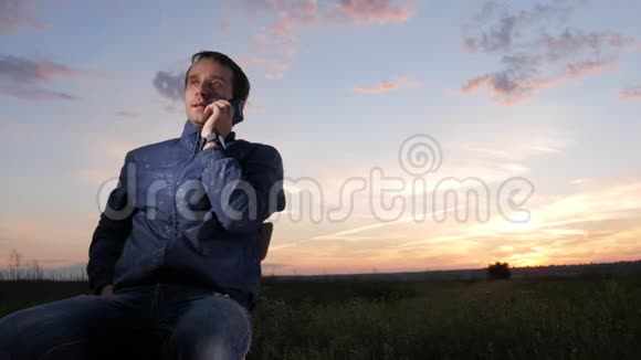 一个年轻人坐在夕阳下用手机说话后面是美丽的天空视频的预览图