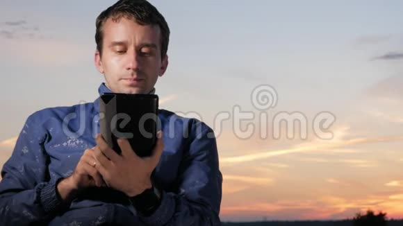 一个年轻人坐在日落时分检查留言以检查平板电脑上的留言后面是美丽的天空视频的预览图