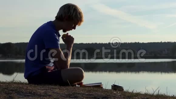 一个年轻的学生在太阳落山时在湖岸读一本书视频的预览图