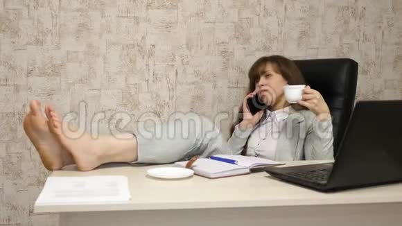 坐在扶手椅上光着脚在桌子上休息喝着茶咖啡的漂亮的女商人工作中的女孩视频的预览图