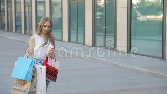 年轻的金发女孩带着幸福的微笑手里拿着许多购物袋喝着咖啡视频的预览图