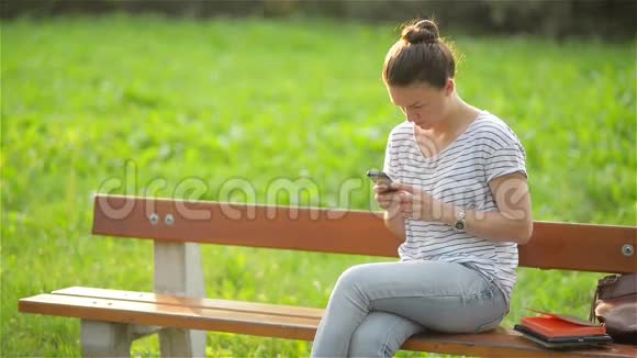 年轻微笑的女人在公园里坐在长凳上用智能手机美丽的欧洲女孩在电话里发短信视频的预览图