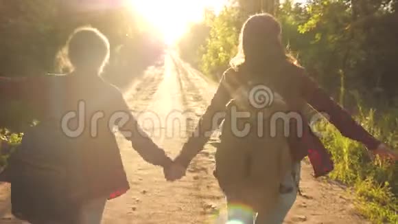 徒步旅行者女孩带着背包的快乐女孩旅行者牵着手在明亮的阳光下沿着乡间的道路奔跑概念视频的预览图
