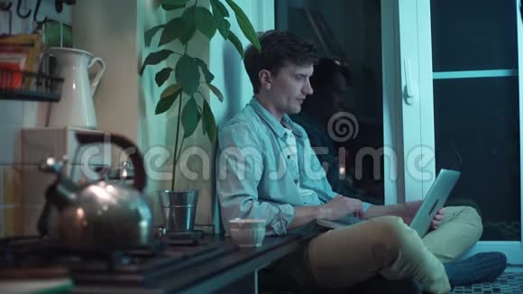 年轻人坐在厨房里用电脑打字而茶壶在炉子上沸腾视频的预览图