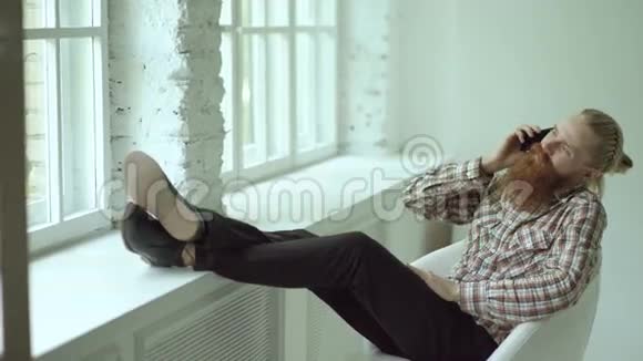 胡子拉碴的年轻时髦商人一边坐在椅子上一边用智能手机说话双腿放在窗台上视频的预览图