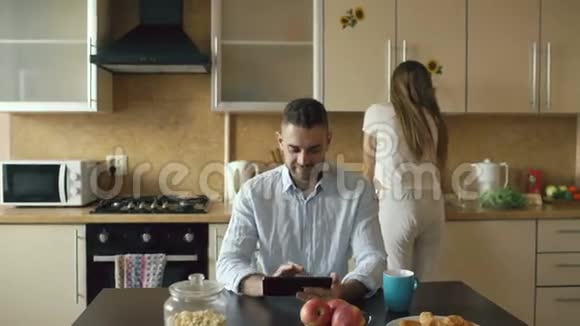 快乐的年轻人坐在厨房里用数字平板电脑而他的女朋友在家做饭视频的预览图