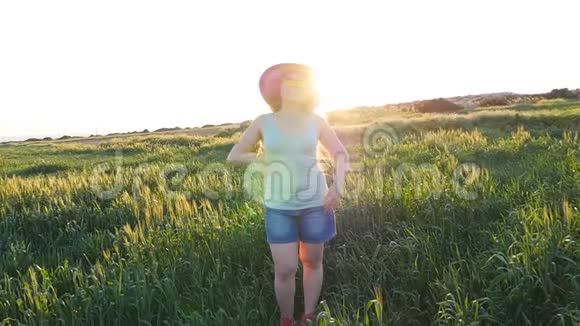 戴帽子的快乐年轻女子在夕阳下的绿野中享受夏日视频的预览图