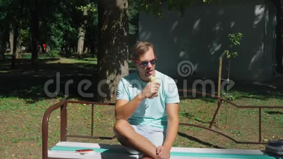 戴墨镜的家伙高兴地吃热狗坐在城市公园的长凳上4k慢动作视频的预览图