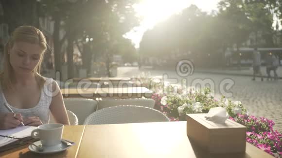 一个穿着白色裙子的年轻金发女孩在咖啡馆外的露台桌上在个人日记中写着思想看着远方视频的预览图