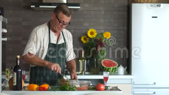 一个戴眼镜的老人在厨房准备晚餐把蔬菜放在碗里搅拌一个有着严肃面孔的养老金领取者视频的预览图