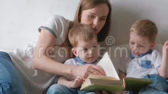 家庭妈妈和两个双胞胎兄弟蹒跚学步的孩子在床上看书家庭阅读时间视频的预览图
