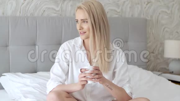 美丽的金发女人在家坐在床上喝咖啡视频的预览图