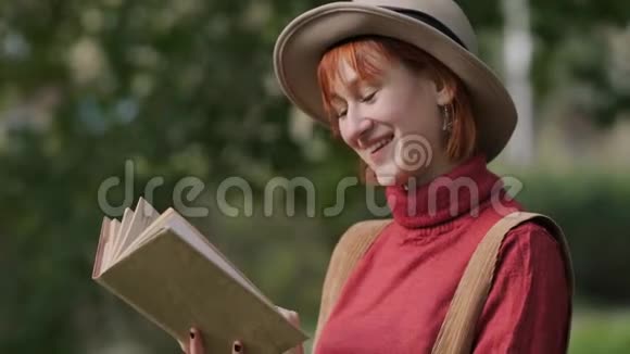 年轻迷人的红头发女人戴着帽子和毛衣在一个自然公园看书秋天凉爽的一天视频的预览图