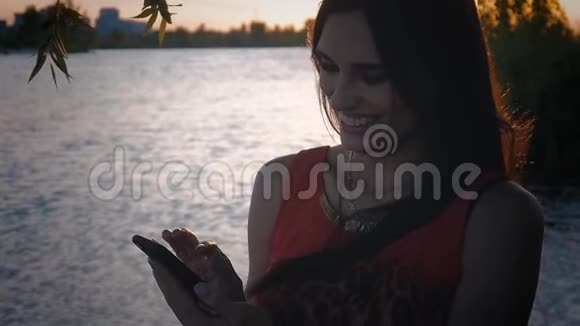 迷人的年轻女性在日落时使用智能手机一个可爱的女孩微笑着在海上使用手机视频的预览图