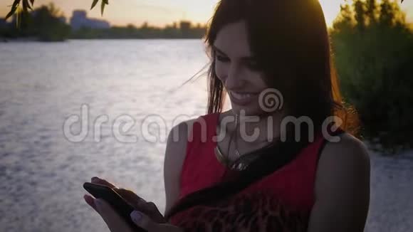 迷人的年轻女性在日落时使用智能手机一个可爱的女孩微笑着在海上使用手机视频的预览图