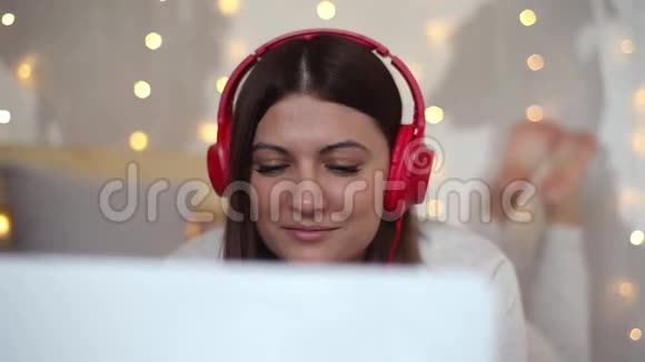 穿着睡衣的漂亮女孩用笔记本电脑在耳机上听音乐心情很好地躺在床上高清高清视频的预览图