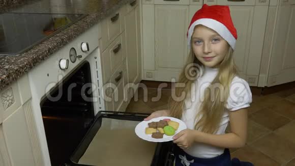 一个年轻的白人女孩把饼干从盘子里扔到烤箱里一个戴红帽子的女孩为圣诞节做准备女孩视频的预览图