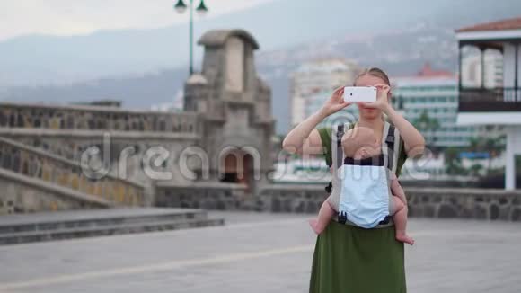 一位年轻的母亲抱着一个婴儿微笑着在她的智能手机上拍照走在街上视频的预览图