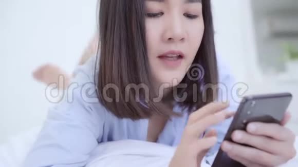 年轻的亚洲女人在早上醒来后躺在床上使用智能手机视频的预览图