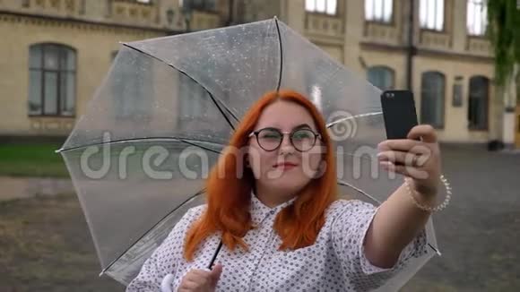 戴眼镜的肥姜女孩正在公园里雨天的智能手机上自拍微笑着撑着伞视频的预览图