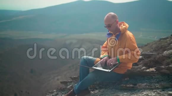 一个穿着黄色夹克蓝色牛仔裤和眼镜的人坐在悬崖边上在笔记本电脑上工作视频的预览图