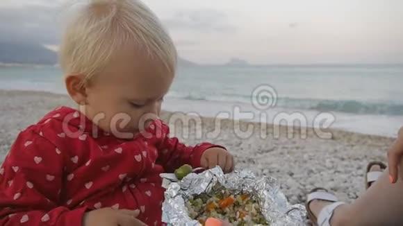 两岁的婴儿在海滩上用铝箔托盘吃绿色橄榄的肖像孩子和妈妈在日落海滩上视频的预览图