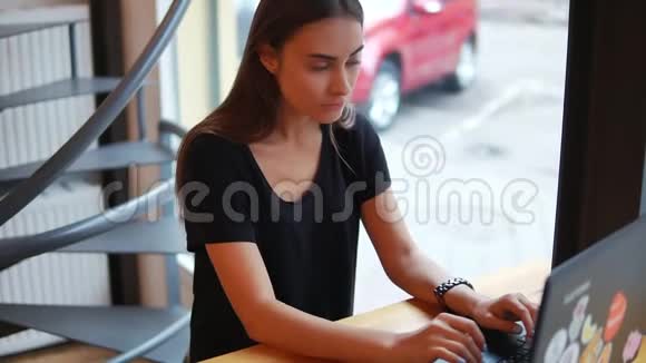 年轻的女人拿着笔记本电脑坐在窗边的咖啡店里想着该写什么或者如何回答然后视频的预览图