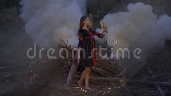 一个带着浅棕色猫头鹰的女巫赤脚站在树林中央的烟雾和发光的动名词之间视频的预览图