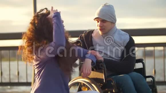 快乐的一对夫妇坐在轮椅上的残疾男子和迷人的年轻女子在夕阳下交谈视频的预览图