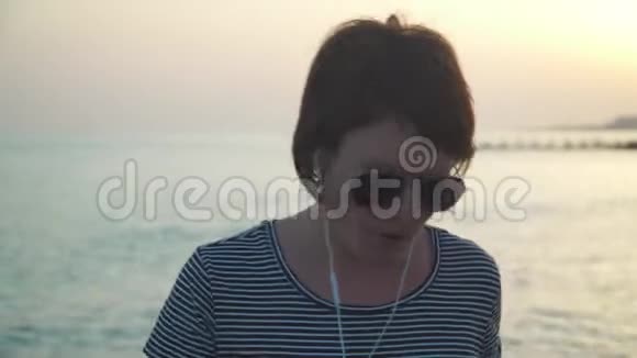 穿着耳机在沙滩上跳舞的闲妇日落时分视频的预览图