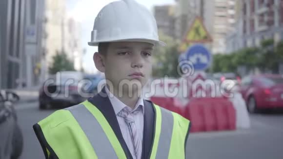 肖像可爱的小男孩穿着商务服安全装备和建筑工人头盔站在繁忙的道路上视频的预览图