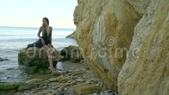 镜头走近一个穿着黑色风衣坐在河口中部石滩上的岩石上的悲伤女孩视频的预览图