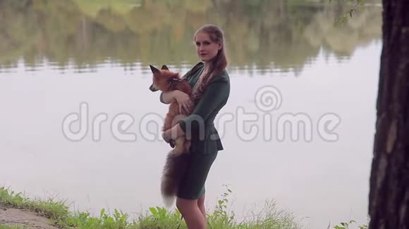 年轻的女人抱着橙色的狐狸夏天站在河岸边视频的预览图