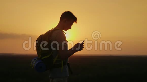 一个带背包的年轻游客在山顶上使用手机在夕阳的余晖中橙色视频的预览图