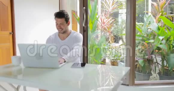 年轻人在家工作手拿电脑坐在桌子上微笑着打字视频的预览图