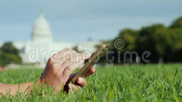 手拿智能手机在草坪上在华盛顿国会大厦的背景下在美国旅行和学习视频的预览图
