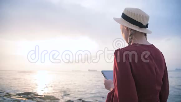 日落时分一个穿着夏红色连衣裙和草帽坐在海边的一块石头上的迷人少女视频的预览图